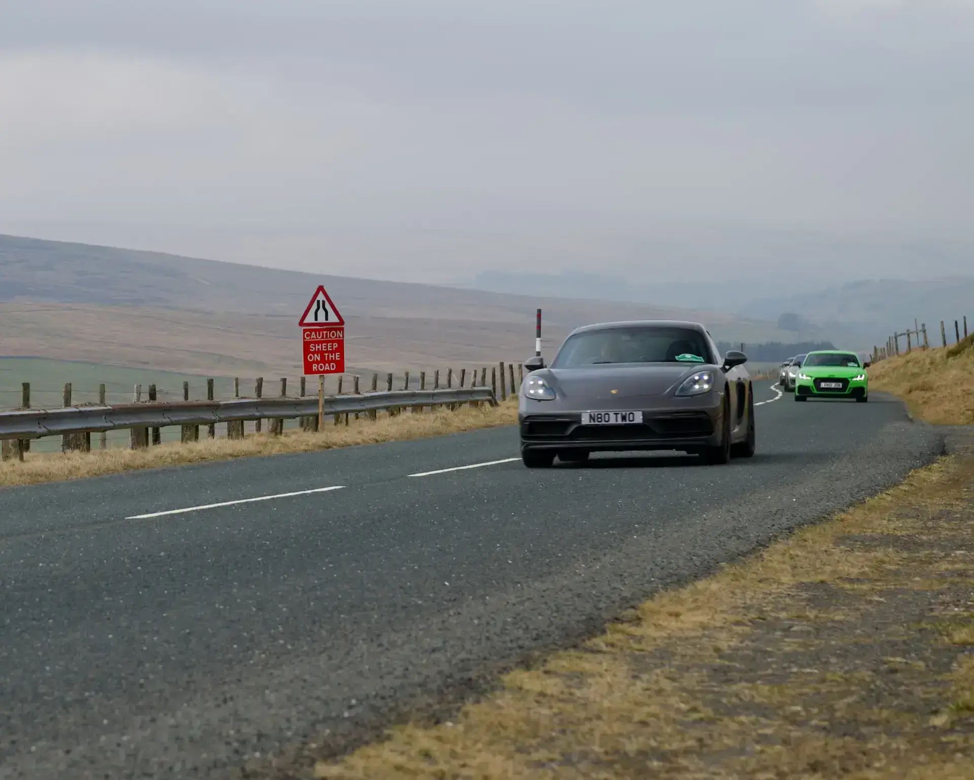 A dark sports car passing a warning sign on a scenic Lake District road.